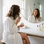 Femme aux cheveux ondulés sans frisottis appliquant une crème coiffante devant un miroir de salle de bain